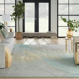 Light grey rug with a soft gradient in blue, beige and grey tones, placed under a sofa in a bright living room with glass doors and pastel decor.