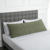 Quilted green body pillow with diamond stitching placed in front of two white pillows on a neatly made bed with a tufted grey headboard.