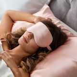 Light pink satin sleep mask embroidered with "COCOON" placed on a matching pink satin pillowcase. Light pink satin sleep mask embroidered with "COCOON" placed on a matching pink satin pillowcase.
