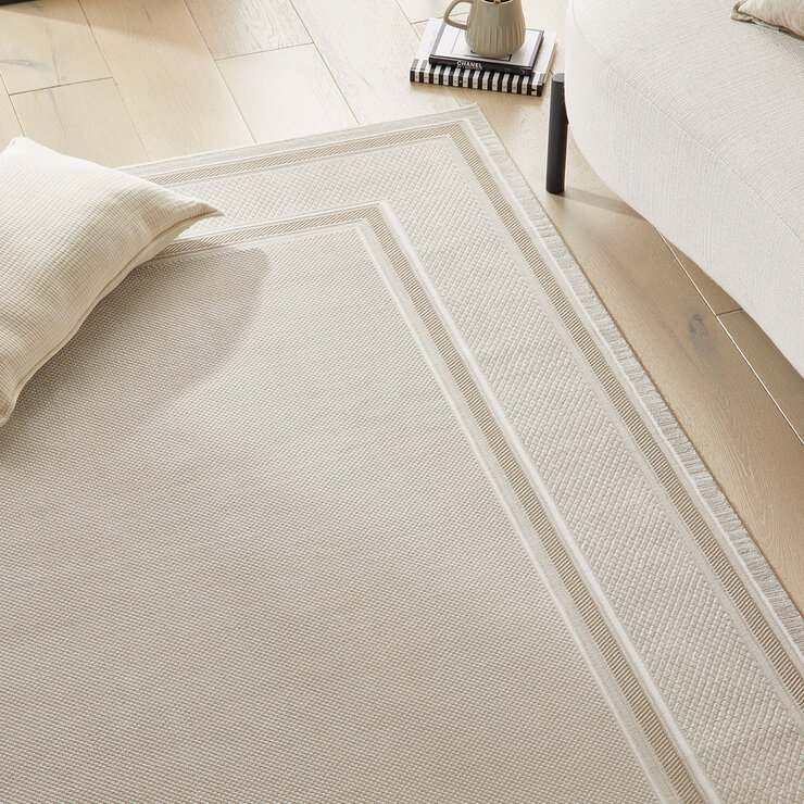 Textured beige rug with a decorative border, shown in a minimalist interior with a pillow, books and soft natural lighting.