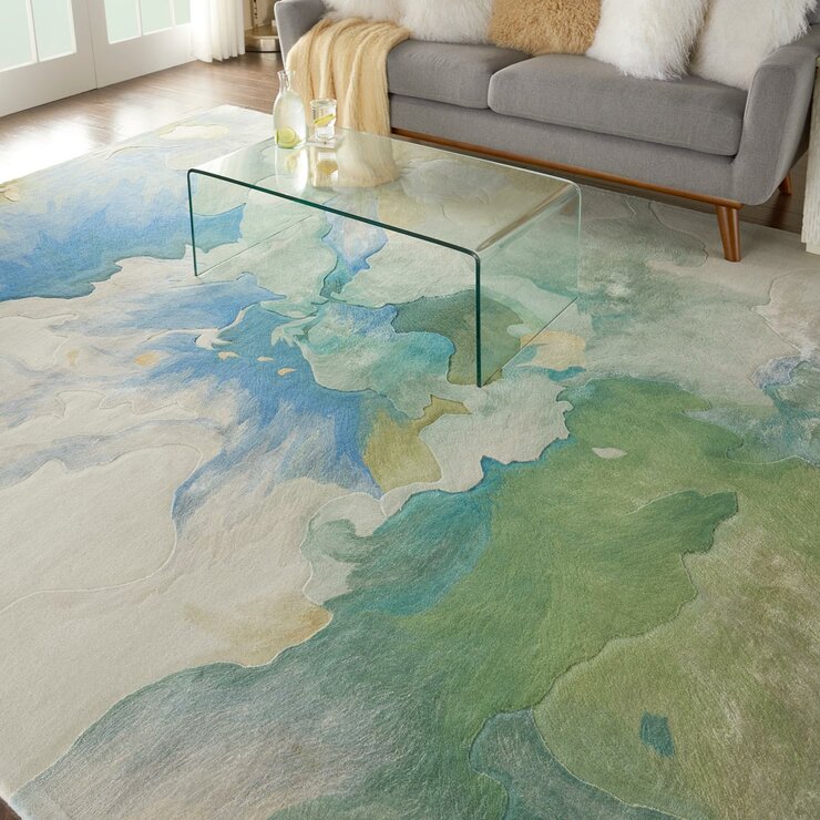Large rug with a flowing design in green, blue, and beige. Placed under a clear glass coffee table in a bright living room.