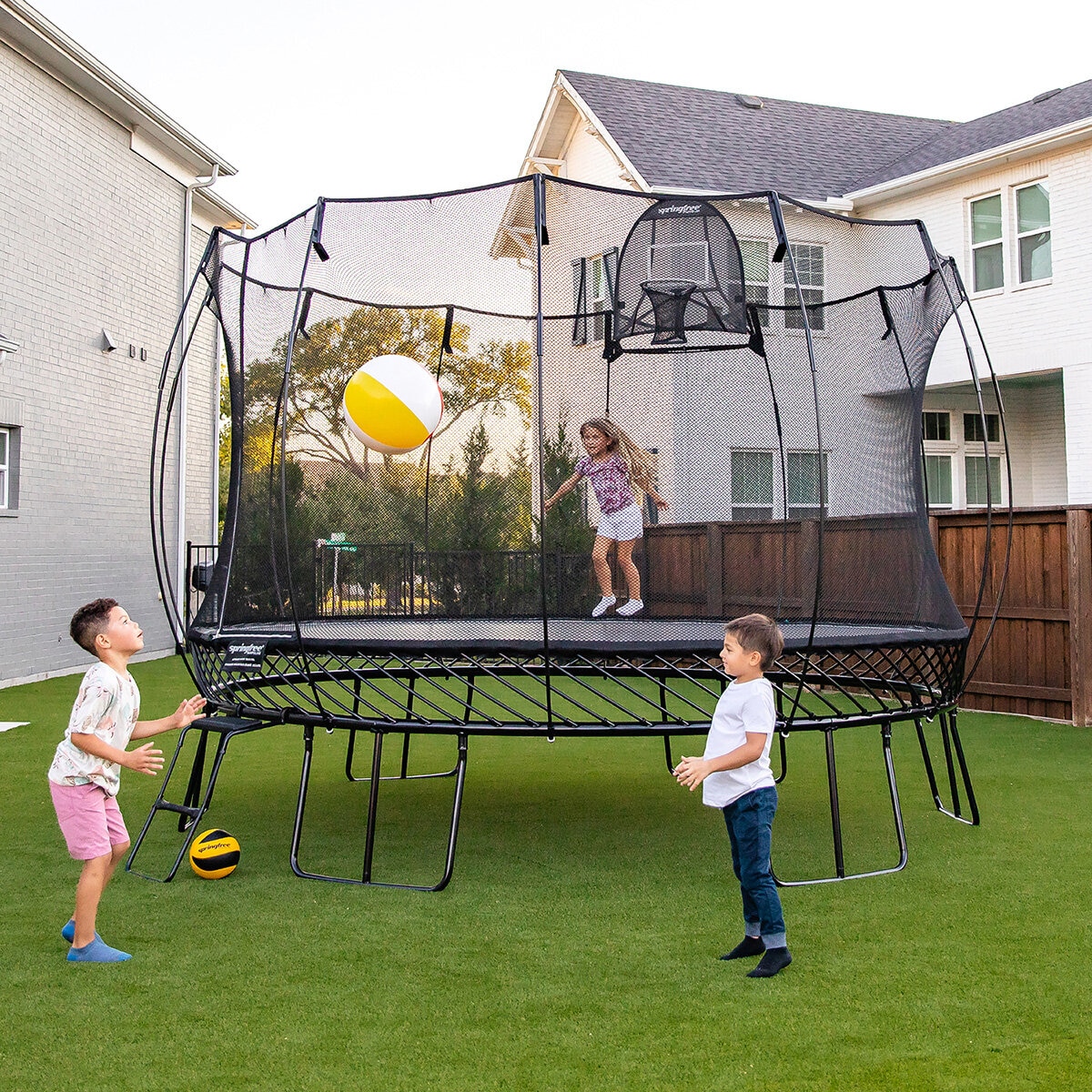 Lifestyle image of a child jumping on the trampoline, and two children playing with a ball beside it Lifestyle image of a child jumping on the trampoline, and two children playing with a ball beside it