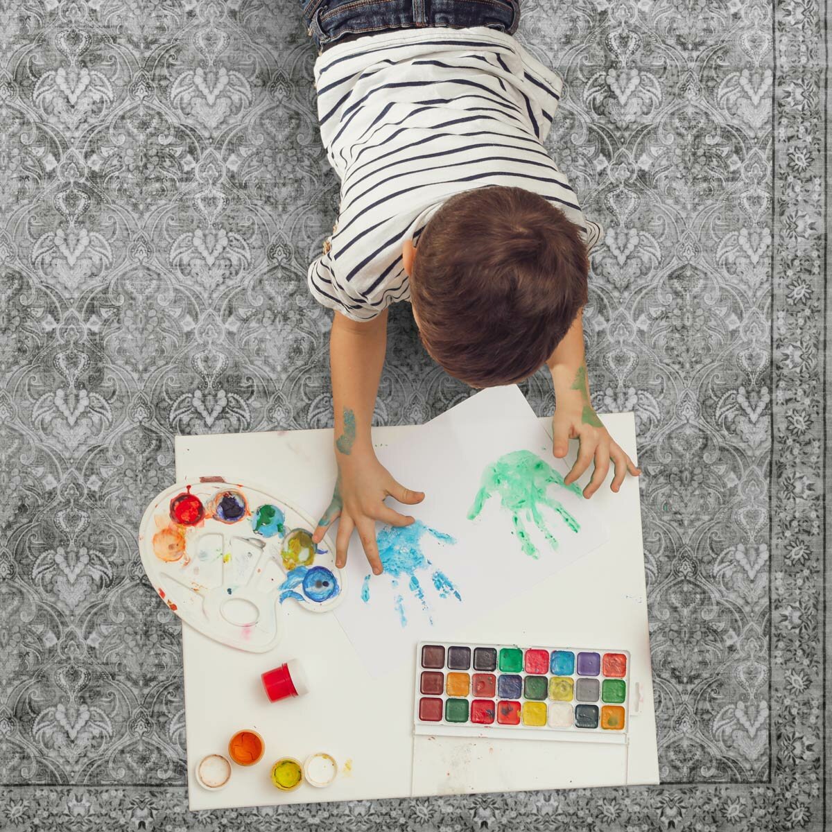 Child friendly green patterned rug used as a play surface, with child painting handprints and art supplies nearby