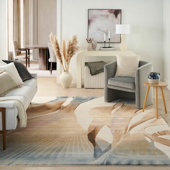 Large rug with an abstract pattern in beige, brown and blue, placed in a modern living room with neutral furniture and soft textures.