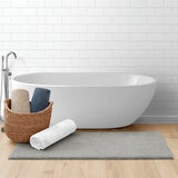 Minimalist bathroom with Grey bath mat laid at the front of a freestanding tub with additional bath mats rolled in shades: Tan,Blue and White