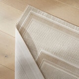 Corner of a beige rug with a raised geometric pattern, flipped to reveal a smooth woven backing on light wood flooring.