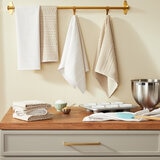 Three neutral-toned kitchen towels hang from a gold rail above a wooden worktop with baking tools