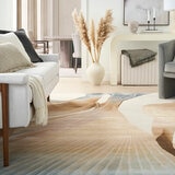 Beige and blue abstract rug laid across a light floor in a bright living room with a white sofa, armchair and pampas grass decor.