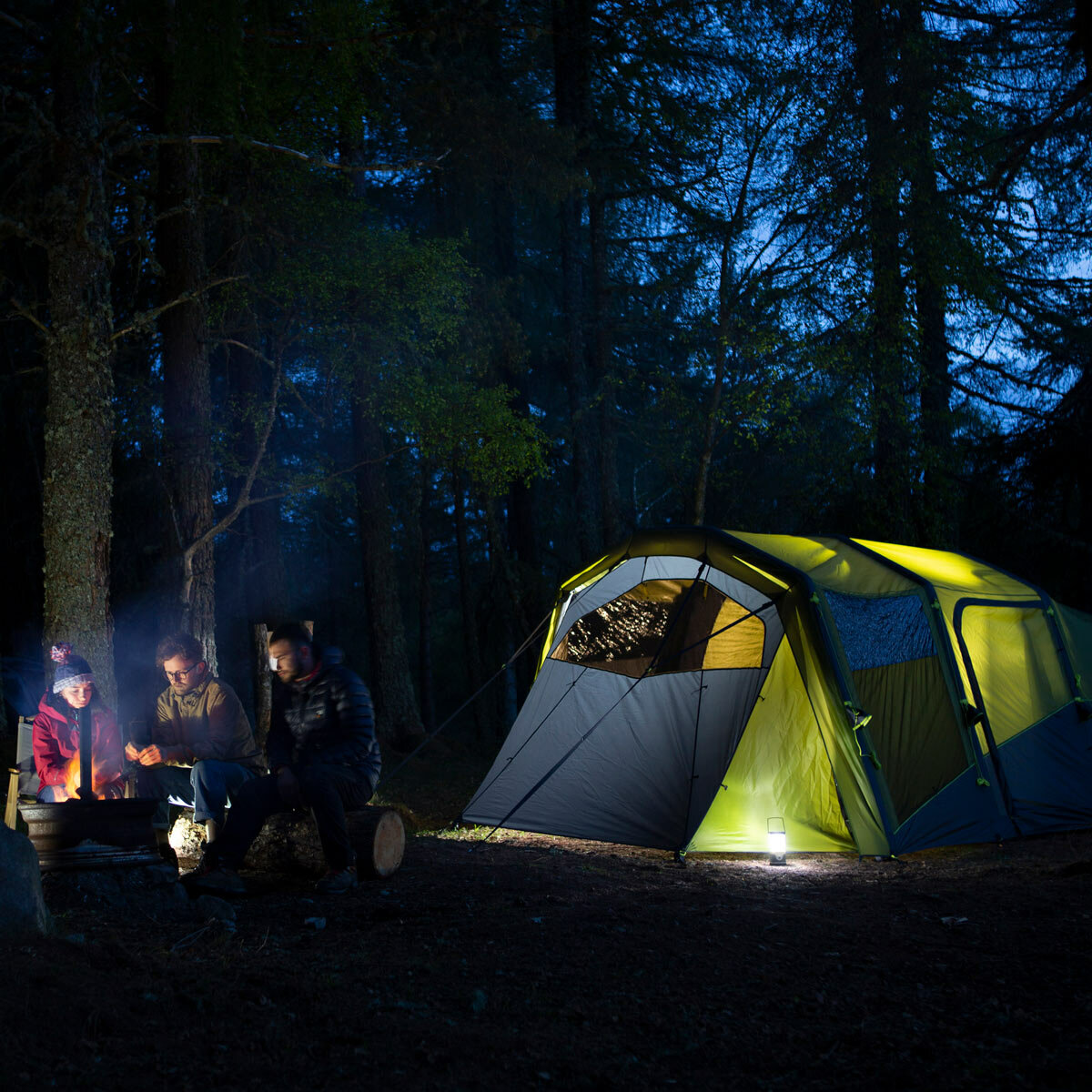 Lifestyle image showing the tent illuminated at night with people sat outside by a campfire Lifestyle image showing the tent illuminated at night with people sat outside by a campfire