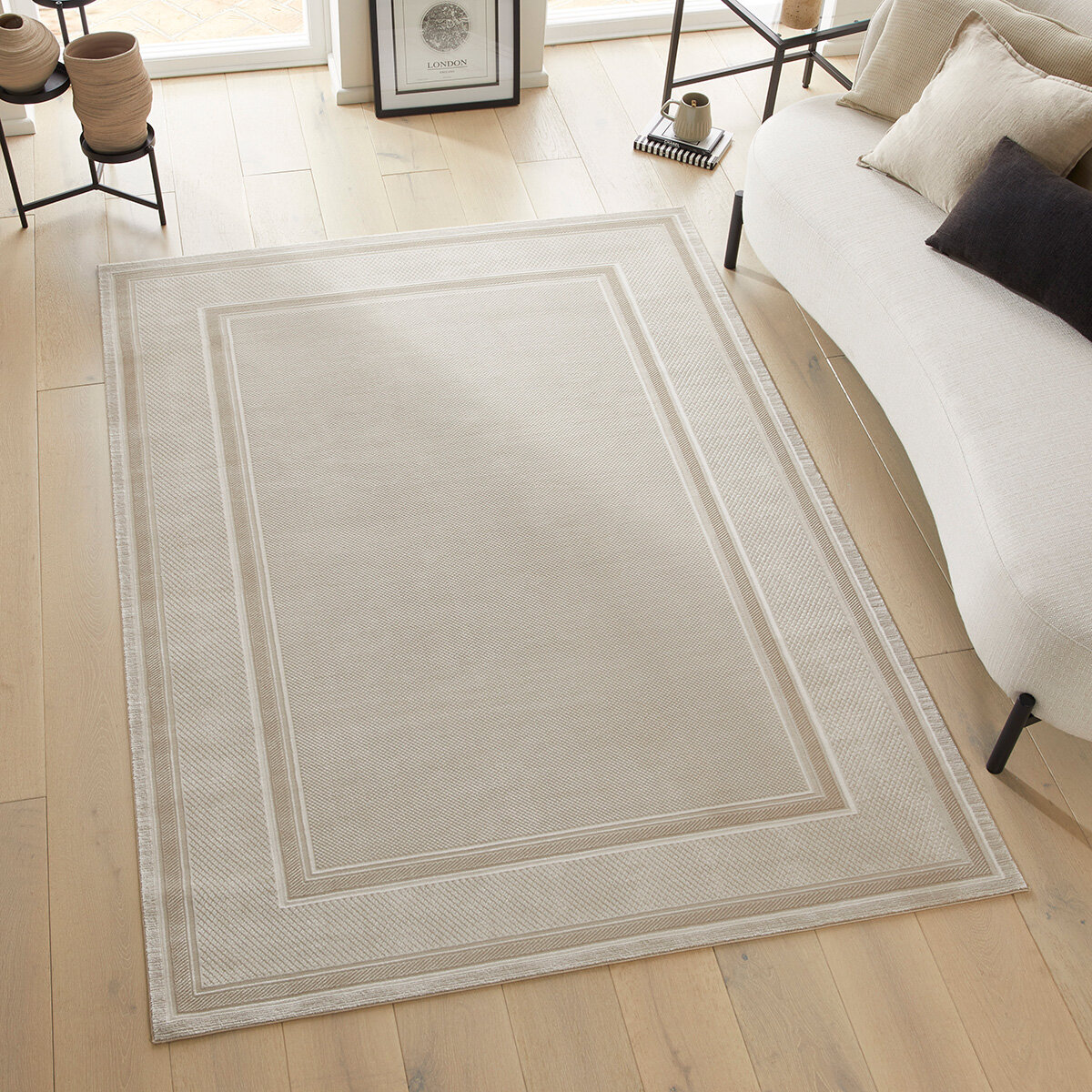Beige rug with layered rectangular borders, styled in a modern living room with light wood flooring and minimal furniture.