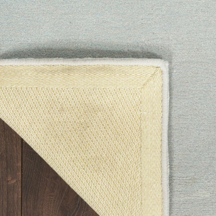 Cream rug with a subtle herringbone texture and white border, shown flipped over to reveal the underside on a pale blue floor. Cream rug with a subtle herringbone texture and white border, shown flipped over to reveal the underside on a pale blue floor.