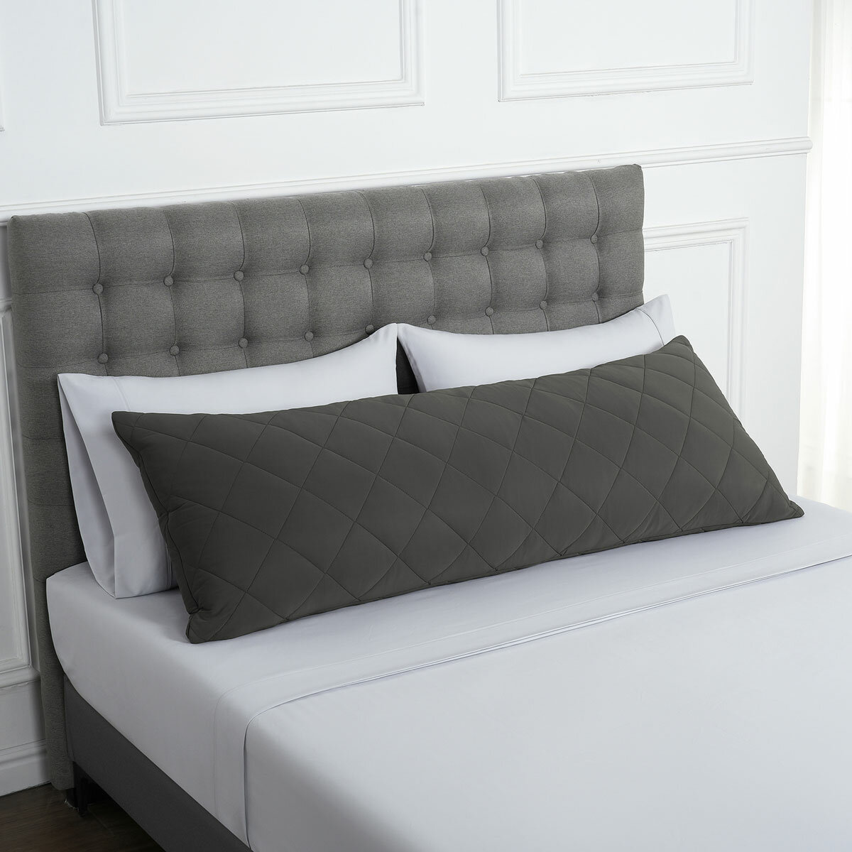 Quilted grey body pillow with diamond stitching placed in front of two white pillows on a neatly made bed with a tufted grey headboard.