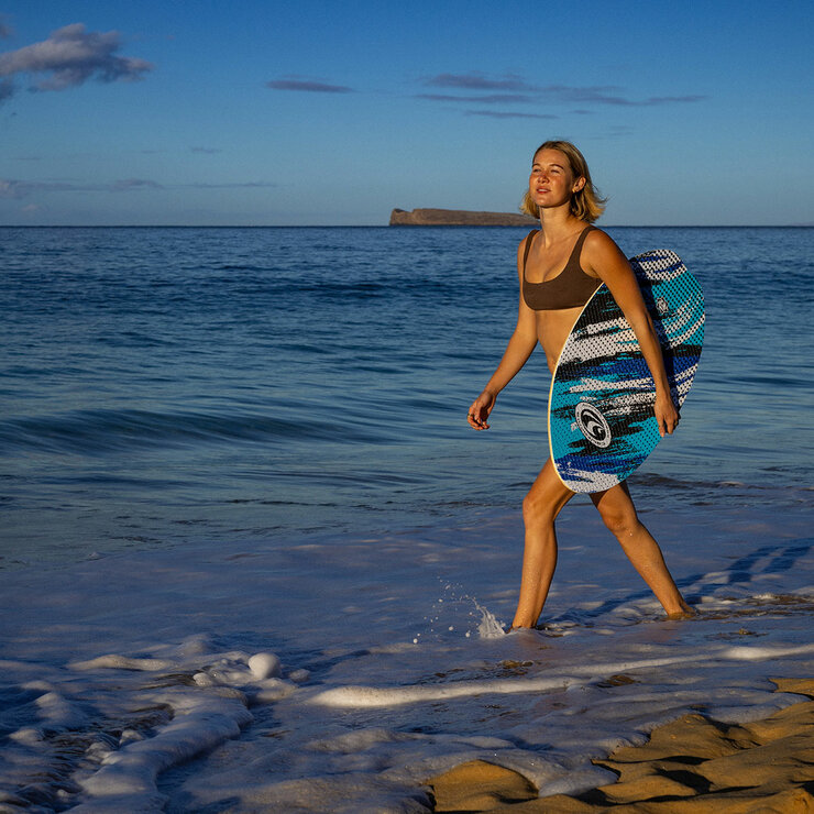 Lifestyle image of a woman walking in the sea holding the skimboard