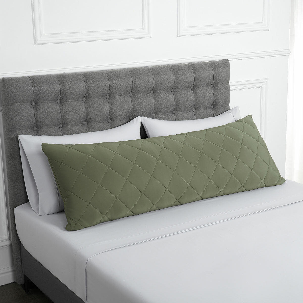 Quilted green body pillow with diamond stitching placed in front of two white pillows on a neatly made bed with a tufted grey headboard.