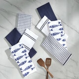Eight navy-themed tea towels arranged on a white marble surface, featuring fish prints, stripes, solids, and grid patterns, with three wooden kitchen utensils below Eight navy-themed tea towels arranged on a white marble surface, featuring fish prints, stripes, solids, and grid patterns, with three wooden kitchen utensils below