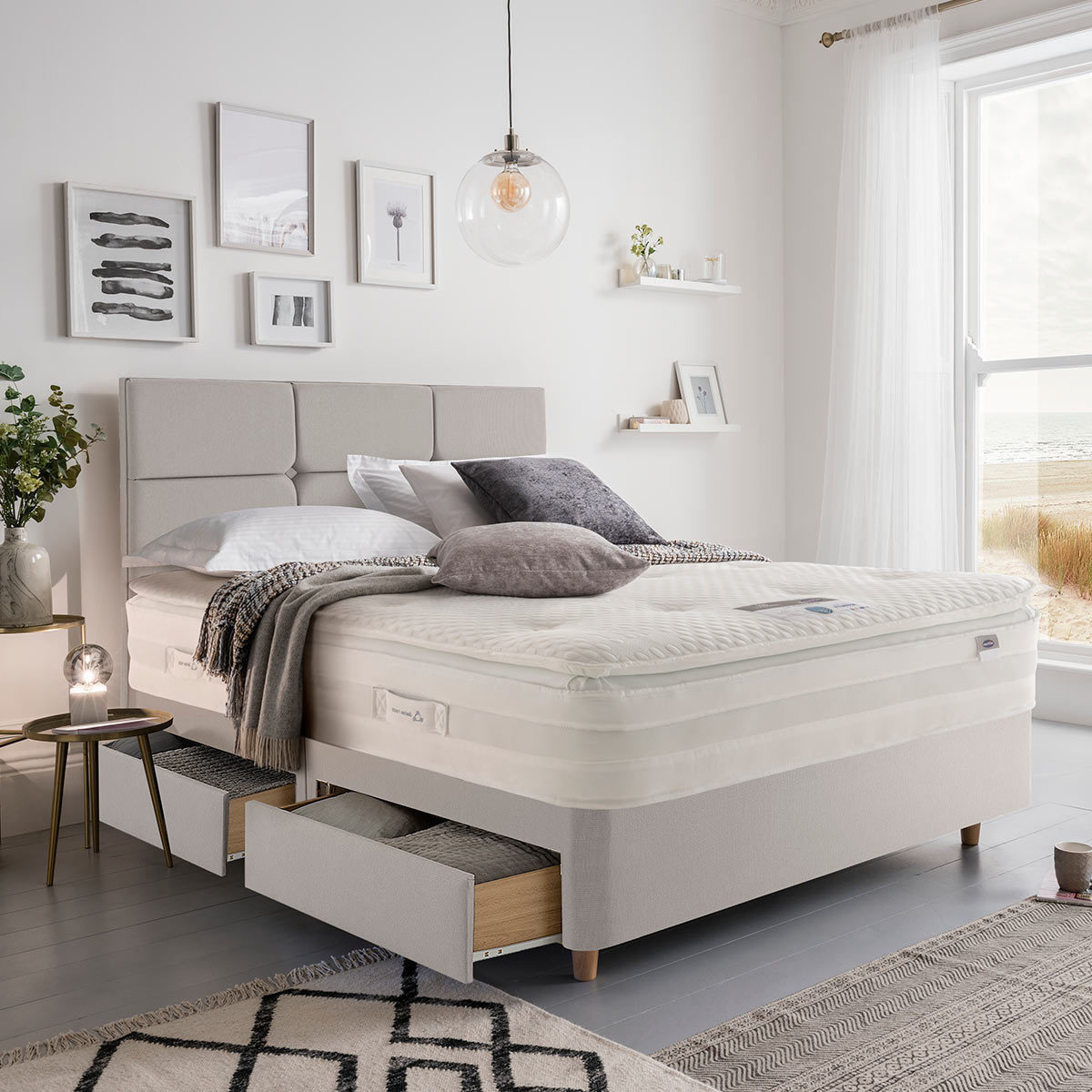 Lifestyle image of made up bed in dove grey in front of window showing beachside