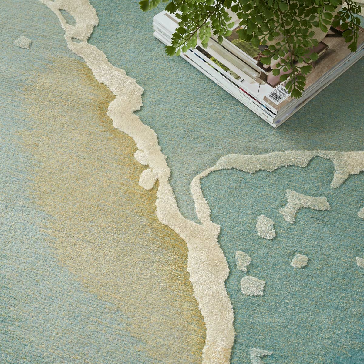 Rug with a beach-inspired design in sandy beige and ocean blue, shown close-up with a stack of magazines and a small plant on top.