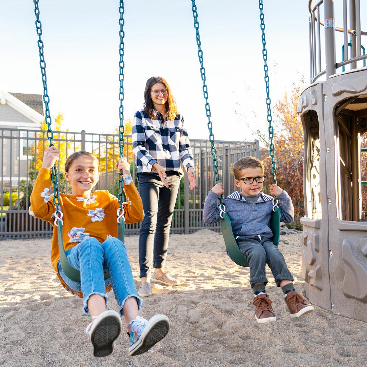 Lifestyle image of a woman pushing two children on the swings Lifestyle image of a woman pushing two children on the swings