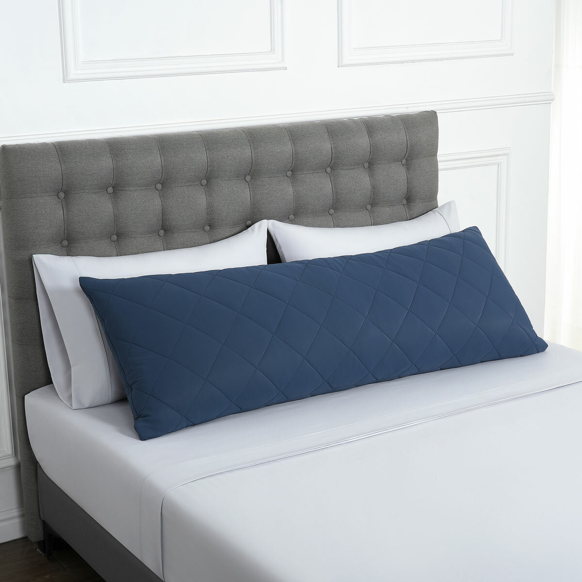 Quilted blue body pillow with diamond stitching placed in front of two white pillows on a neatly made bed with a tufted grey headboard.